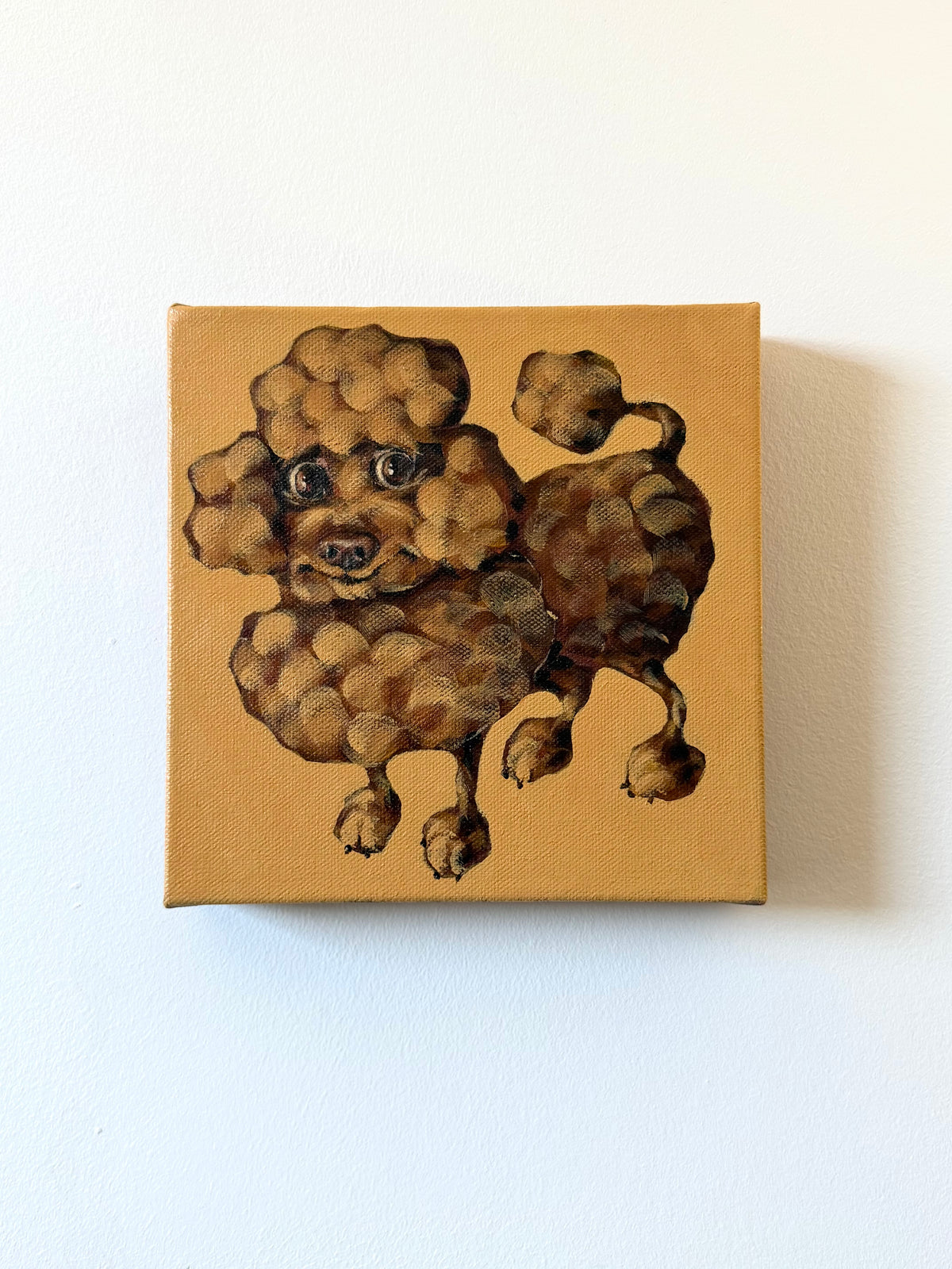 A square canvas painting of a curly-haired poodle on a warm tan background, mounted on a plain white wall.