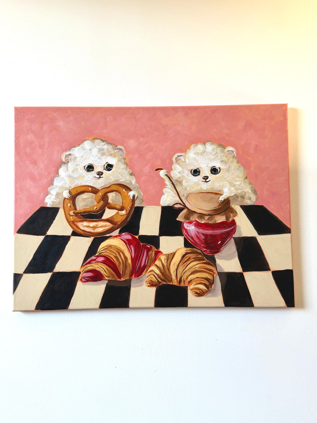 A square canvas painting depicting two fluffy white dogs sitting at a black-and-white checkered table, eating croissants, a pretzel, and coffee with a mug. The background of the painting is pink, and the canvas is displayed against a plain white wall.