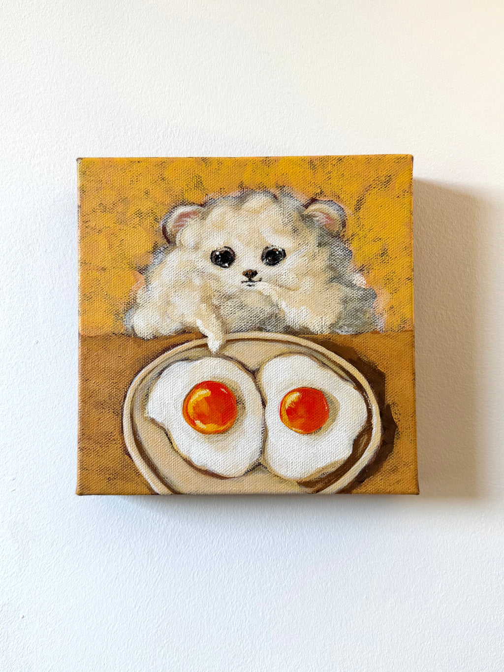 A small square canvas painting hanging on a white wall, depicting a fluffy cream-colored hamster peeking over a plate of two fried eggs with bright orange yolks.