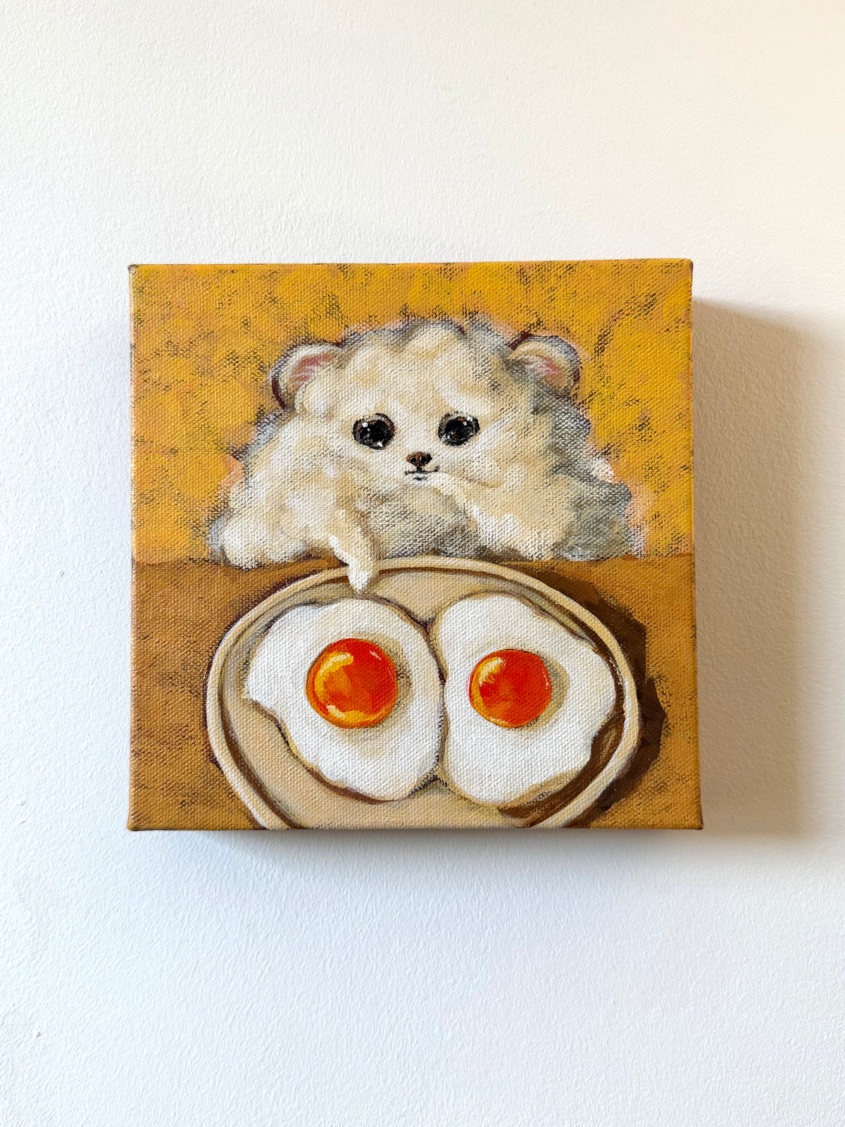 A small square canvas painting hanging on a white wall, depicting a fluffy cream-colored hamster peeking over a plate of two fried eggs with bright orange yolks.