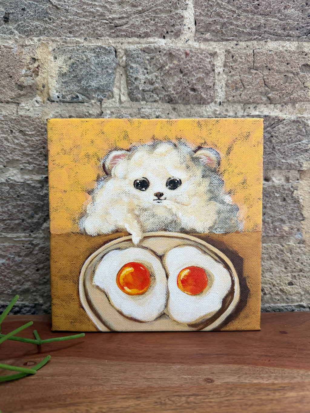 A square canvas painting of a fluffy cream-colored puppy peeking over two fried eggs on a wooden table, set against a rustic gray brick wall background.