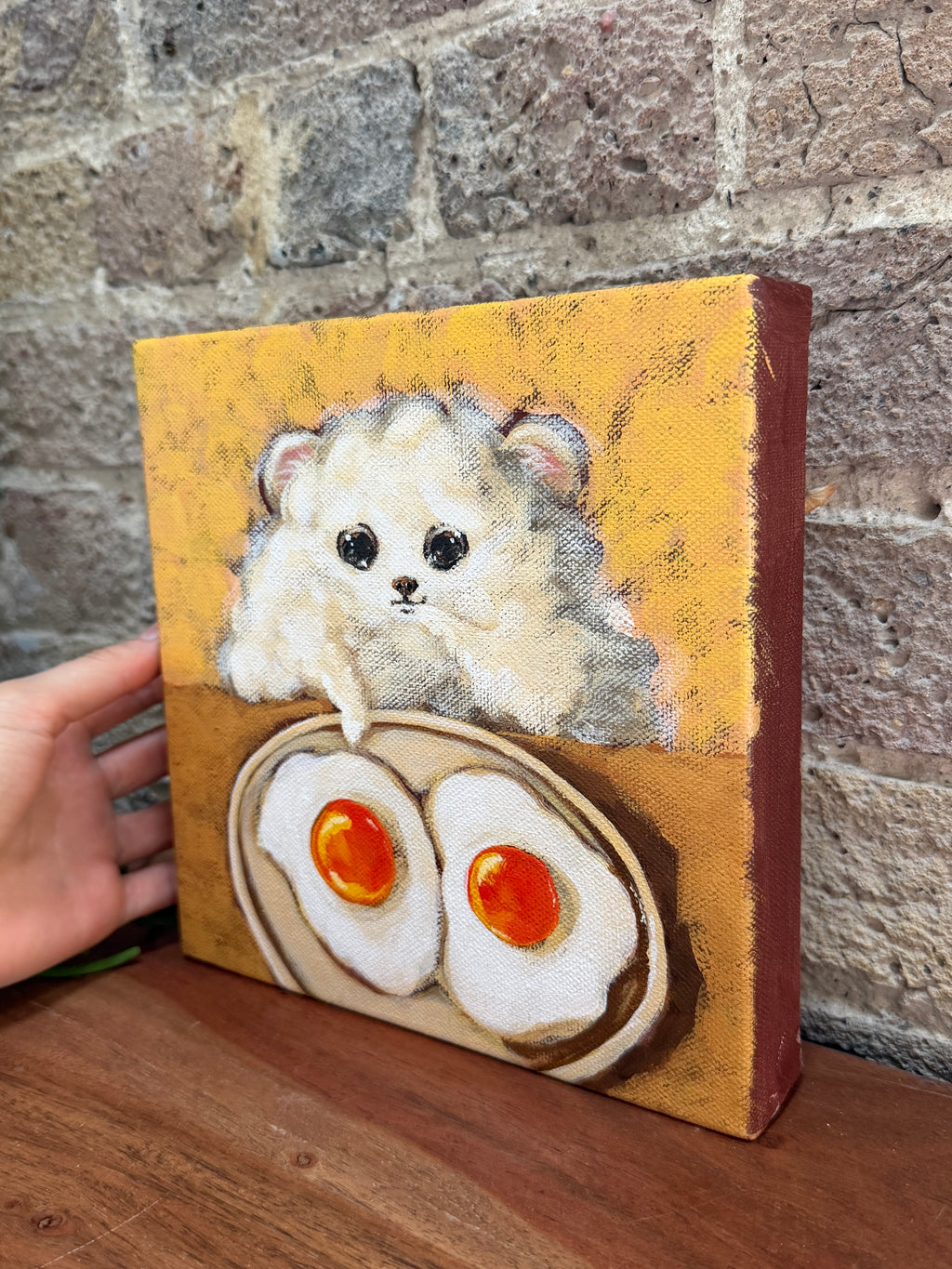 A hand holds a small canvas painting featuring a fluffy white hamster above two fried eggs on a yellow-orange background, set against a rustic brick wall and wooden surface.