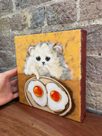 A hand holds a small canvas painting featuring a fluffy white hamster above two fried eggs on a yellow-orange background, set against a rustic brick wall and wooden surface.