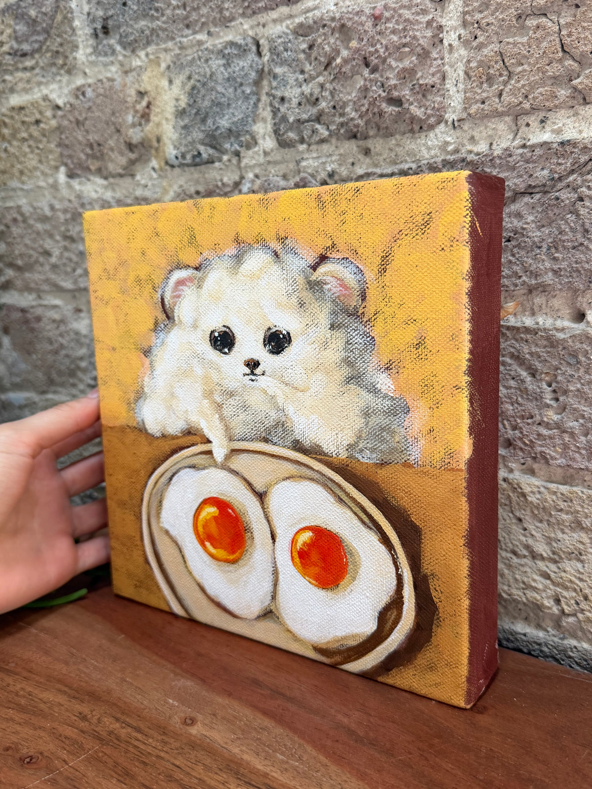 A hand holds a small canvas painting featuring a fluffy white hamster above two fried eggs on a yellow-orange background, set against a rustic brick wall and wooden surface.