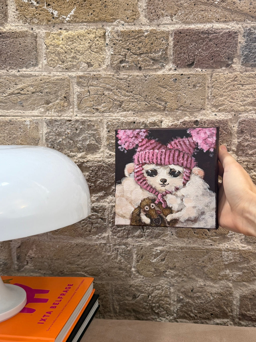 A hand holds a square canvas print featuring a fluffy white cat wearing a pink knit hat and scarf against a dark background, positioned in front of a rustic brick wall with a white table lamp and orange books partially visible.