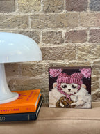 A square framed art print depicting a white dog in a pink knit hat and scarf, styled on a beige sofa next to a white table lamp and books against a brick wall.