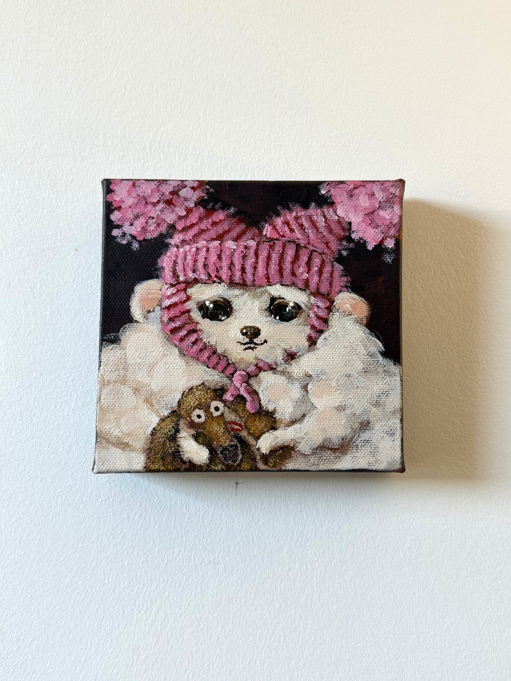 A square canvas painting hanging on a white wall, depicting a white fluffy dog wearing a pink striped knit hat and scarf, with pink flowers in the background and a small brown animal at its feet.