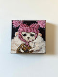 A square canvas painting hanging on a white wall, depicting a white fluffy dog wearing a pink striped knit hat and scarf, with pink flowers in the background and a small brown animal at its feet.