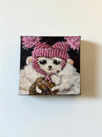 A square canvas painting hanging on a white wall, depicting a white fluffy dog wearing a pink striped knit hat and scarf, with pink flowers in the background and a small brown animal at its feet.