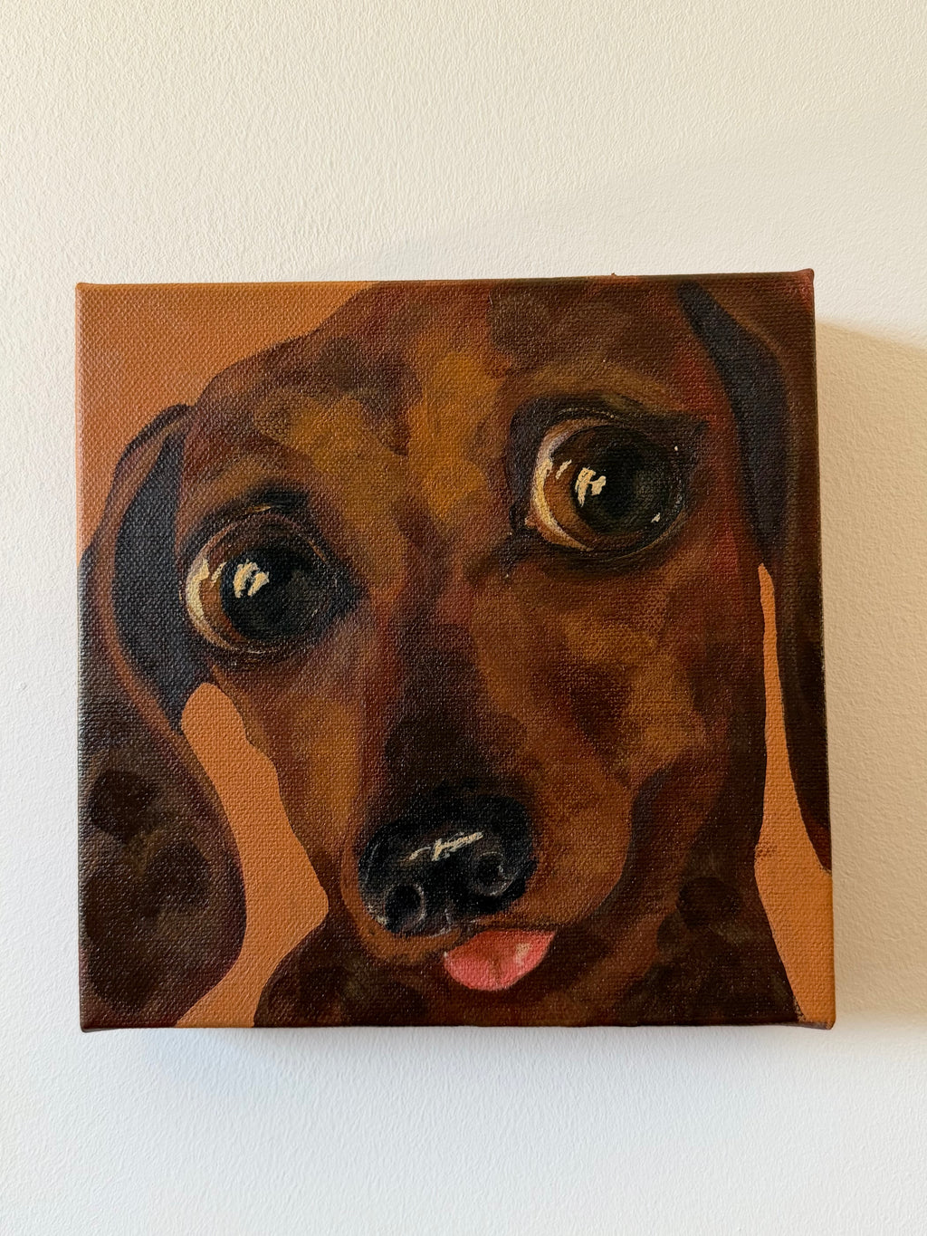 A square canvas painting of a dachshund dog's face, shown hanging on a plain off-white wall. The painting features warm brown and tan tones with dark brown shading and a small pink tongue visible.