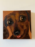 A square canvas painting of a dachshund dog's face, shown hanging on a plain off-white wall. The painting features warm brown and tan tones with dark brown shading and a small pink tongue visible.