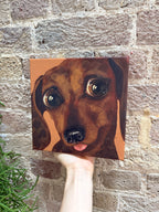 A hand holds a square canvas painting of a dachshund dog's face against a rustic brick wall background.