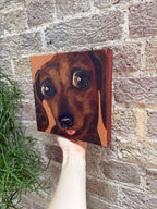 A hand holds a square canvas painting of a dachshund dog's face against a brick wall background.