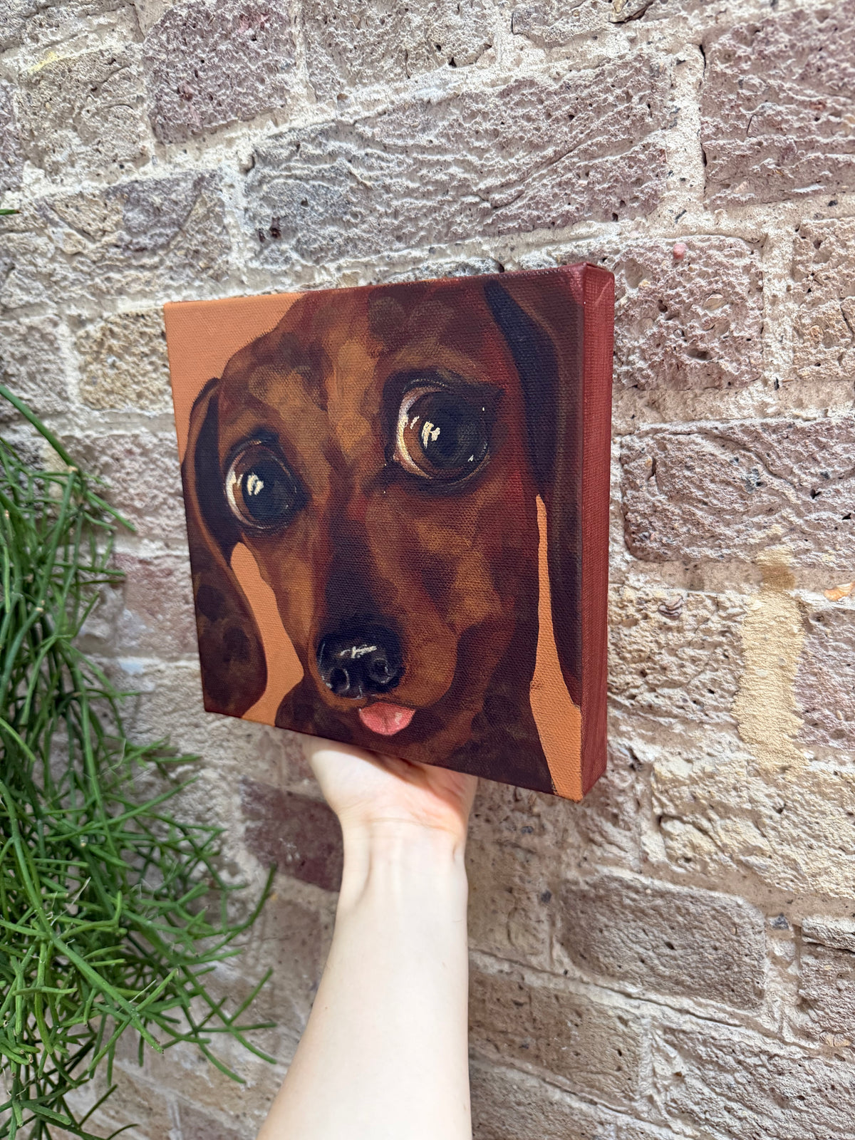 A hand holds a square canvas painting of a dachshund dog's face against a brick wall background.