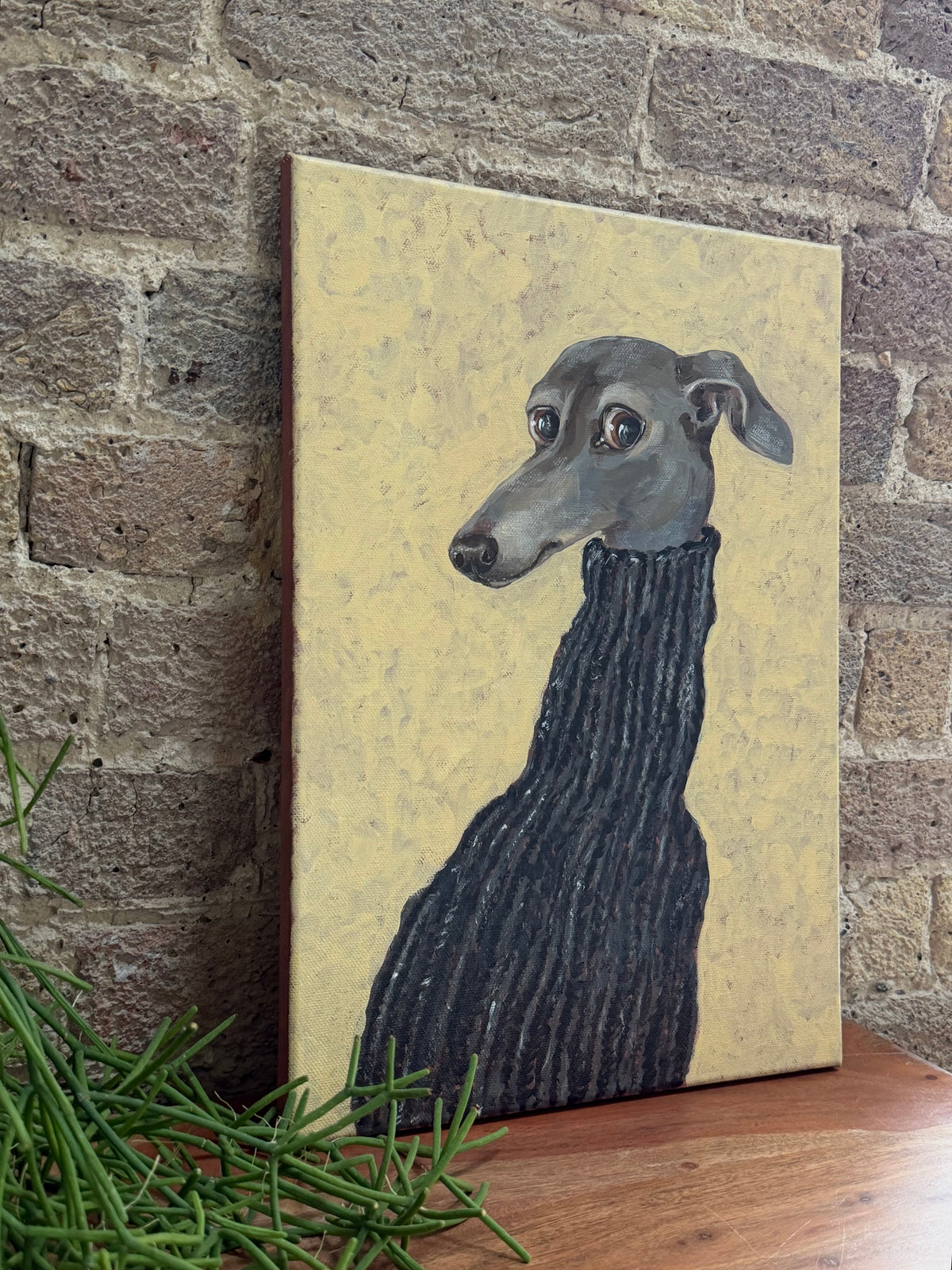 A vertical canvas painting of a greyhound dog wearing a dark ribbed sweater, set against a textured beige background, leaning against a rustic brick wall on a wooden surface with green foliage in the foreground.