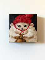 A square canvas painting of a fluffy cream-colored dog wearing a bright red knit hat and red bow tie, set against a dark background, mounted on a plain off-white wall.