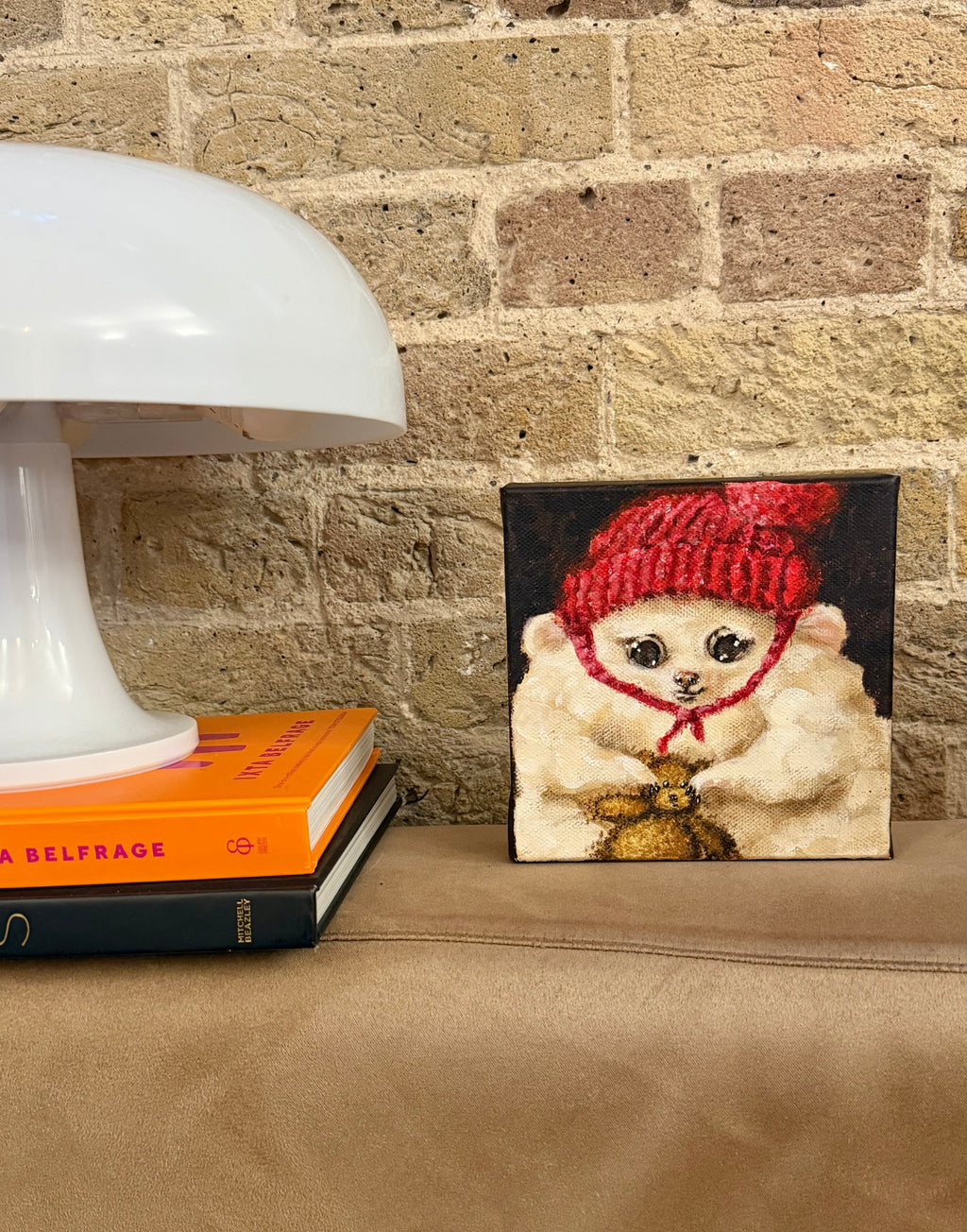 A small square canvas painting of a fluffy cream-colored cat wearing a red knit hat and a gold bow tie, displayed on a tan leather surface next to a white table lamp and books against a brick wall.
