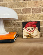 A small square canvas painting of a fluffy cream-colored cat wearing a red knit hat and a gold bow tie, displayed on a tan leather surface next to a white table lamp and books against a brick wall.