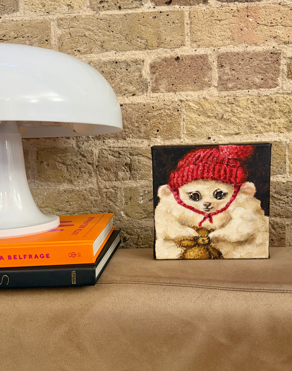 A small square canvas painting of a fluffy cream-colored cat wearing a red knit hat and a gold bow tie, displayed on a tan leather surface next to a white table lamp and books against a brick wall.