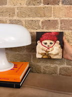 A hand holds a small square canvas painting of a fluffy cream-colored cat wearing a red knit hat and red bow tie against a dark background, set against a brick wall next to a white table lamp and books.